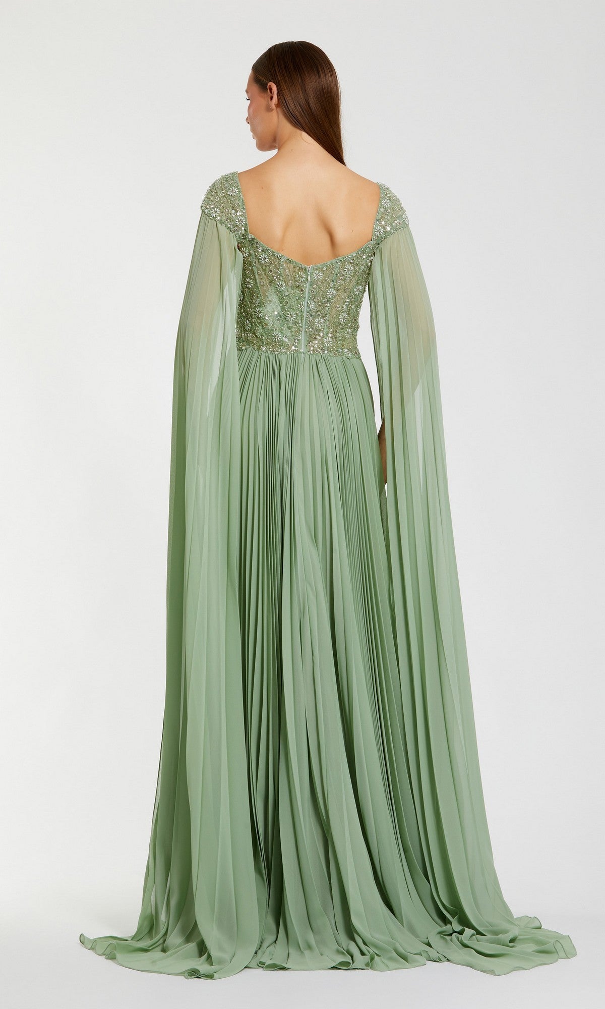 A woman is seen from the back wearing the Mac Duggal 37034 long formal dress, a floor-length light green gown with a floral embellished bodice and long, flowing cape sleeves.